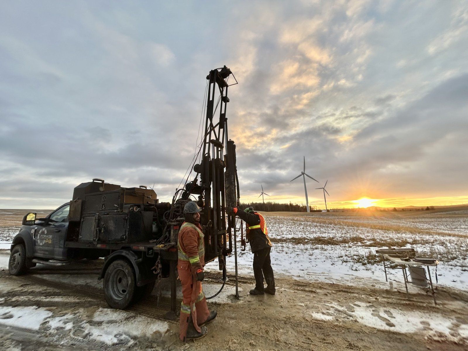 Deux techniciens à la foreuse pendant un forage stratigraphique, dans un champ enneigé bordé d’éoliennes